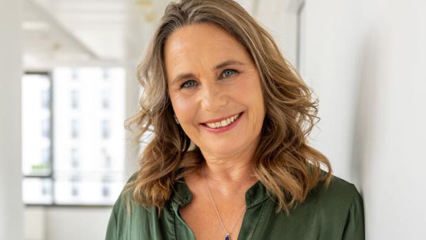 Frau mit schulterlangen, welligen Haaren und grünem Oberteil lächelt in einem hellen, modernen Raum.