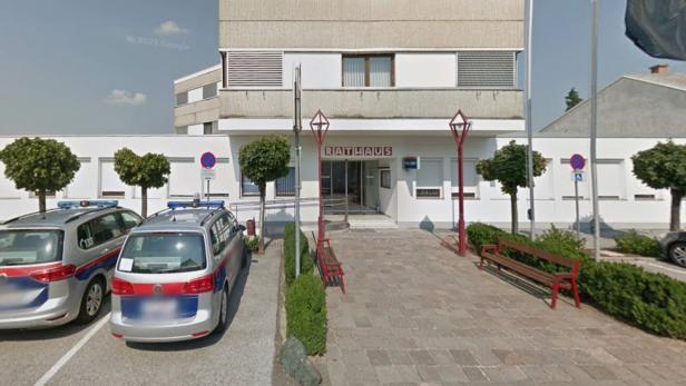 Vor dem Eingang eines Rathauses stehen zwei Polizeiautos, Bänke und kleine Bäume säumen den Weg.