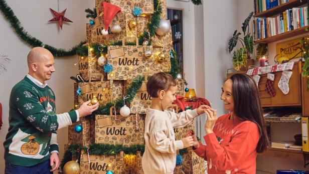 Eine Familie mit einem Kind sitzt um einen Christbaum, der aus Papiersackerl gebastelt ist, und überreicht sich Geschenke. 