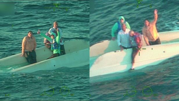 Vier Männer warten auf einem gekenterten Katamaran im offenen Meer auf Rettung und winken einem Hubschrauber der Küstenwache zu.