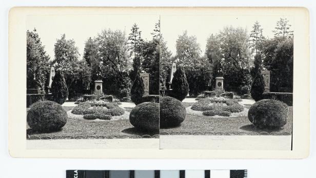Symmetrisch angelegter Garten mit runden Sträuchern, Blumenbeeten, Denkmälern und Bäumen im Hintergrund.