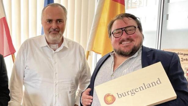 Zwei Männer stehen nebeneinander, einer hält eine Geschenkbox mit der Aufschrift „Burgenland“, im Hintergrund sind Flaggen zu sehen.