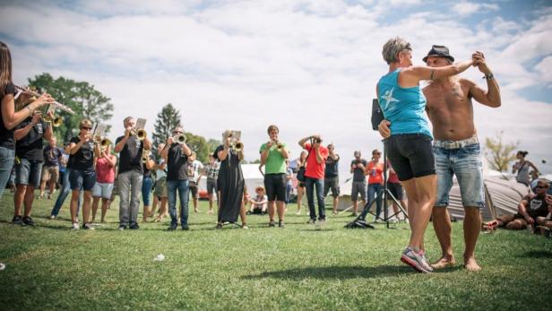Ein Paar tanzt barfuß auf einer Wiese, während eine Blaskapelle im Hintergrund spielt und Menschen zuschauen.
