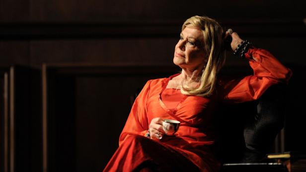 Ingrid van Bergen in einem roten Kleid sitzt mit einer Tasse in der Hand und blickt nach oben.