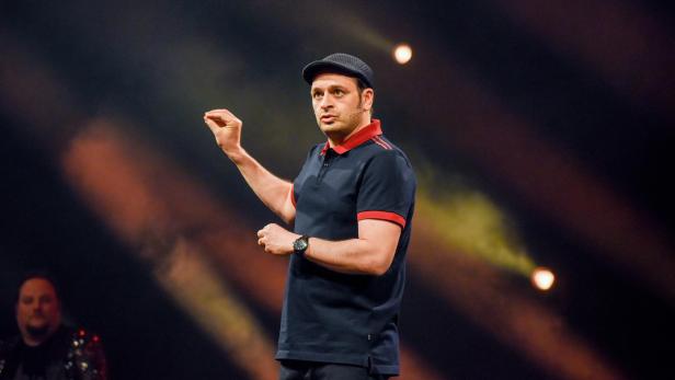 Kaya Yanar mit Schirmmütze und Poloshirt gestikuliert auf einer Bühne.