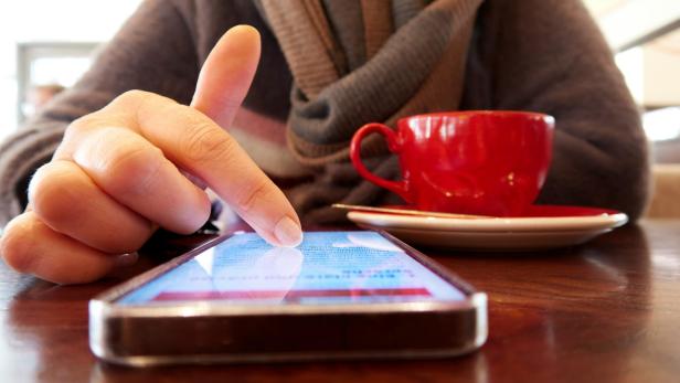 Eine Person scrollt auf einem Smartphone, während eine rote Tasse auf dem Tisch steht.