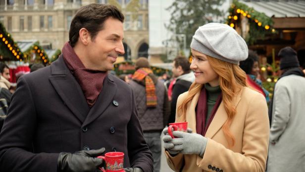 Ein Mann und eine Frau unterhalten sich lächelnd auf einem Weihnachtsmarkt und halten rote Tassen in den Händen.