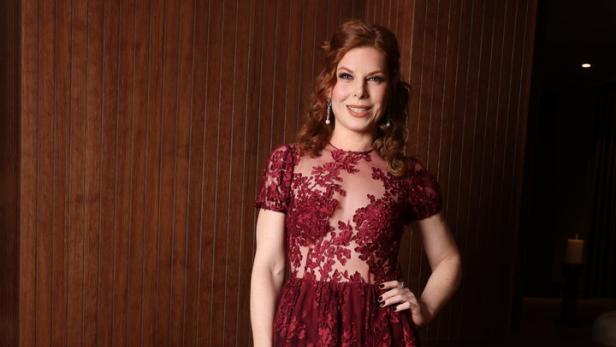 Frau mit langen, gelockten Haaren trägt ein bodenlanges, bordeauxrotes Spitzenkleid mit floralen Mustern.