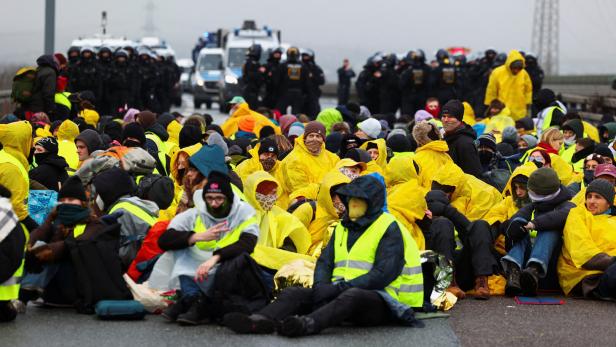 Eine Gruppe Demonstranten in gelben Regenmänteln sitzt auf einer Straße, während Polizisten im Hintergrund stehen.