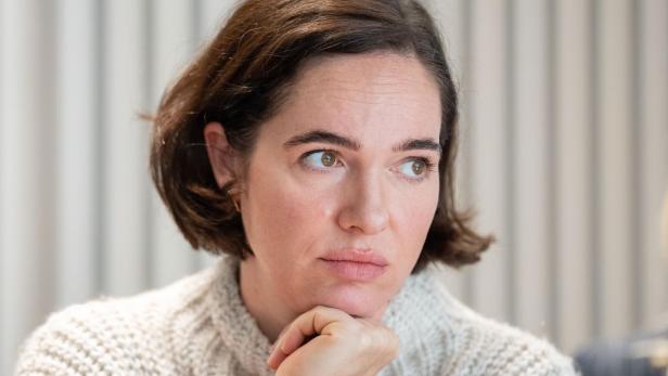 Verena Altenberger mit braunen Haaren und einem hellen Pullover stützt ihr Kinn nachdenklich ab.