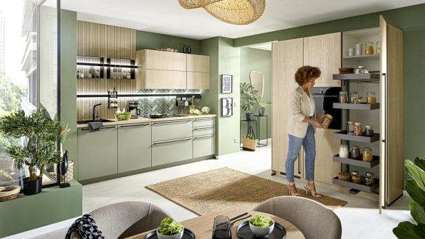 Moderne Küche mit grünen und Holzelementen, eine Frau nimmt ein Glas aus einem offenen Vorratsschrank.