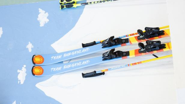 Zwei Paar Skier mit der Aufschrift „Team Burgenland“ und zwei Skistöcke liegen nebeneinander auf einer bemalten Fläche.