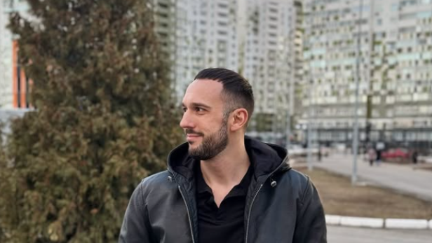 Mann steht in schwarzer Jacke und schwarzen Hosen, darüber einer Lederjacke, mit den Händen in den Hosentaschen vor einem Tannenbaum. 