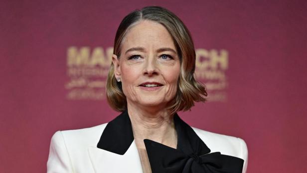 Jodie Foster trägt einen weißen Blazer mit schwarzem Revers und einer großen schwarzen Schleife vor einem roten Hintergrund.