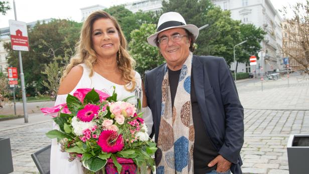 Eine Frau im weißen Kleid hält einen bunten Blumenstrauß, neben ihr steht ein Mann mit Hut und Schal, beide lächeln.