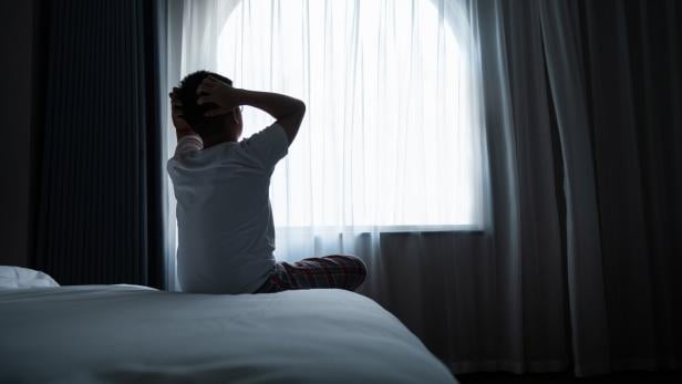 Ein Mann sitzt auf einem Bett und blickt auf ein helles, rundes Fenster.