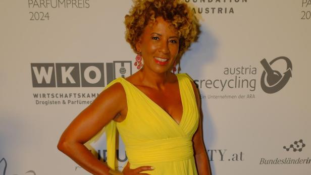 Eine Frau mit lockigen Haaren trägt ein langes, gelbes Kleid und steht lächelnd vor einer Sponsorenwand.