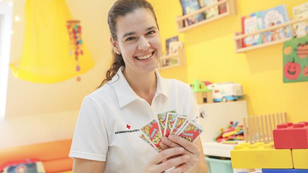 Eine lächelnde Frau in weißem Poloshirt hält Uno-Junior-Karten in einem bunt eingerichteten Kinderzimmer.