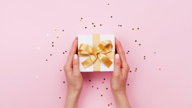 Zwei Hände halten eine weiße Geschenkbox mit goldener Schleife auf rosa Hintergrund mit kleinen Sternkonfetti.