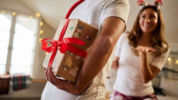 Ein Mann versteckt ein Geschenk mit roter Schleife hinter dem Rücken, während eine Frau erwartungsvoll lächelt.