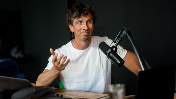 Mann mit weißem T-Shirt gestikuliert mit einer Hand vor einem Mikrofon an einem Tisch mit Laptop und Glas.