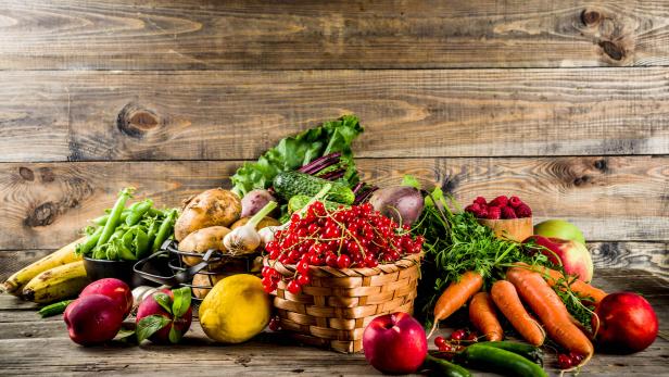 Korb mit roten Johannisbeeren, umgeben von Karotten, Äpfeln, Kartoffeln, Gurken, Bananen, Erbsen und anderem Gemüse auf Holztisch.