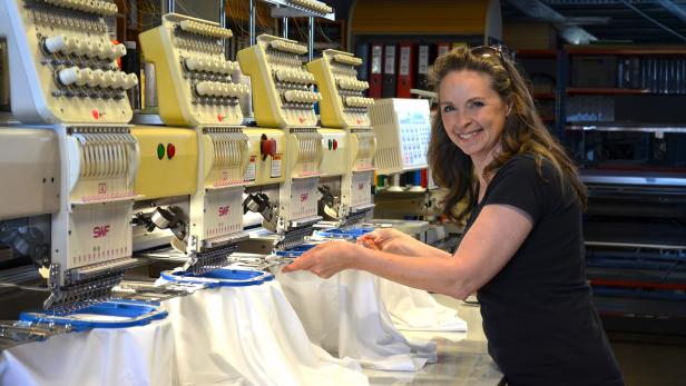 Frau mit dunklem T-Shirt bedient mehrere industrielle Stickmaschinen mit weißen Stoffen in einer Werkstatt.