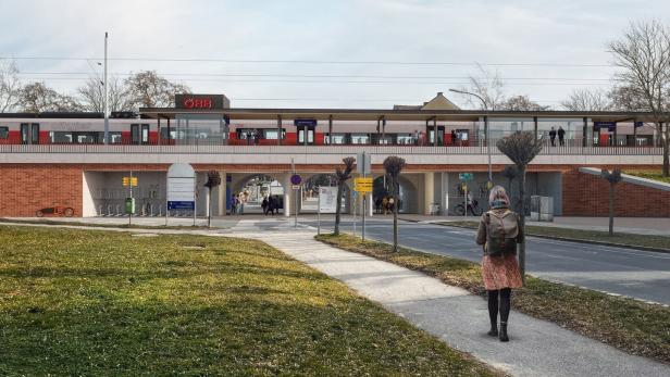 Eine Person mit Rucksack geht auf einen modernen Bahnhof mit rotem Zug und mehreren Passanten zu.
