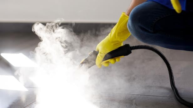 Hand mit gelbem Handschuh reinigt Bodenfliesen mit einem Dampfreiniger.