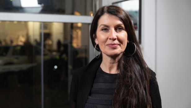 Frau mit langen dunklen Haaren und großen Ohrringen steht lächelnd in einem modernen Innenraum mit Glasfront.