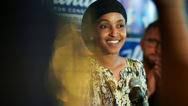 Ilhan Omar lächelt vor einem Mikrofon, im Hintergrund ist ein Wahlplakat zu sehen.