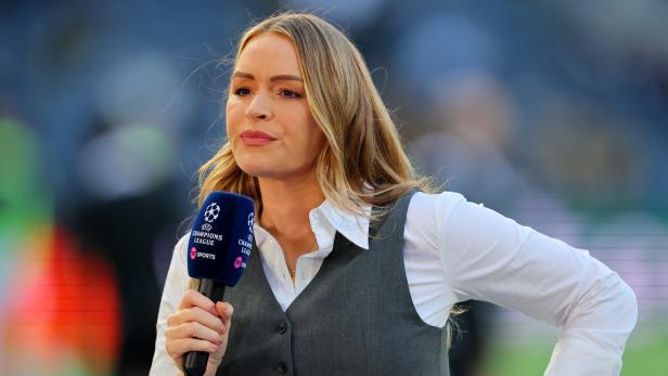 Eine Frau mit langen blonden Haaren hält ein Mikrofon mit Champions-League-Logo und spricht auf einem Sportplatz.