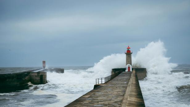 Wellen schlagen gegen einen Leuchtturm am Ende eines langen, gepflasterten Hafendamms unter bewölktem Himmel.