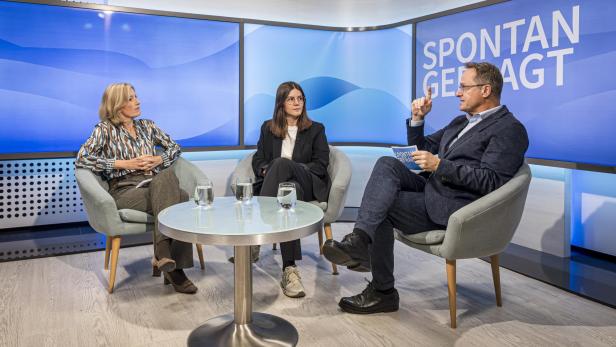 Drei Personen sitzen in Sesseln um einen runden Glastisch mit drei Wassergläsern, im Hintergrund Schriftzug 'SPONTAN GEFRAGT'.