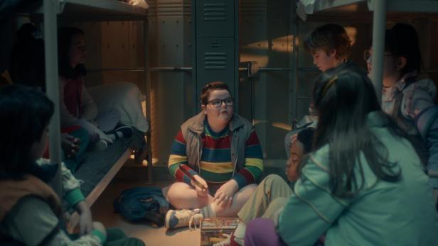 Mehrere Kinder sitzen in einem Schlafsaal im Kreis auf dem Boden zwischen Etagenbetten und unterhalten sich.