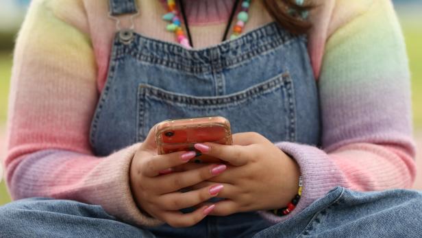 Person in Latzhose hält ein Smartphone mit pink lackierten Nägeln.