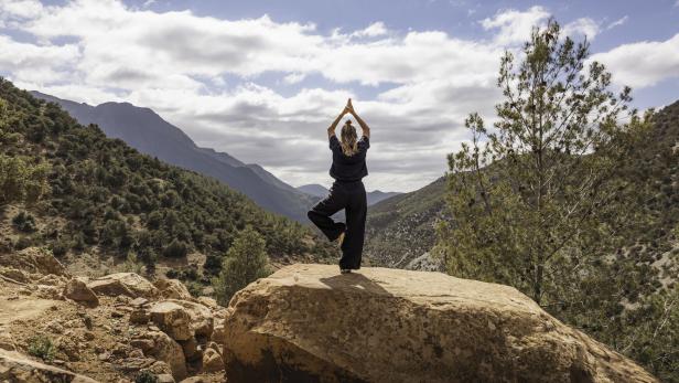 One woman practising yoga in tree pose on rock overlooking forested mountains in nature in the Atlas Mountains, Ourika Valley, Morocco