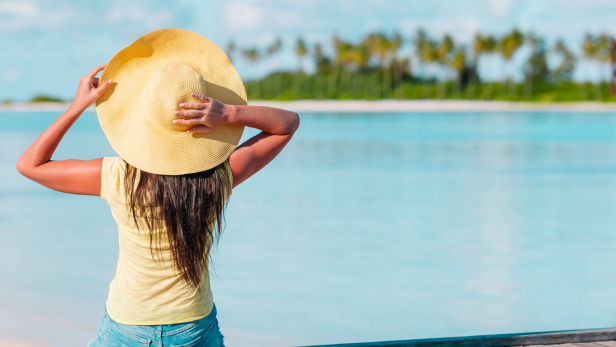 Frau mit Sonnenhut sitzt am Wasser und blickt auf Palmen am gegenüberliegenden Ufer.