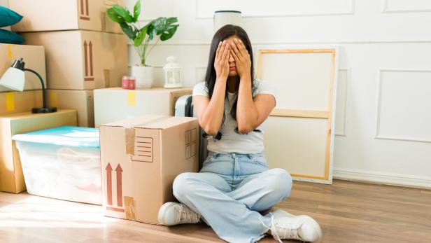 Eine Frau sitzt auf dem Boden zwischen Umzugskartons und bedeckt ihr Gesicht mit den Händen.