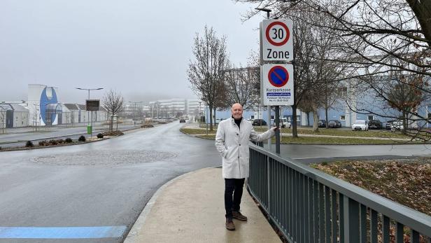 Ein Mann im hellen Mantel steht an einem Geländer neben Verkehrsschildern auf einer nassen, leeren Straße mit kahlen Bäumen.