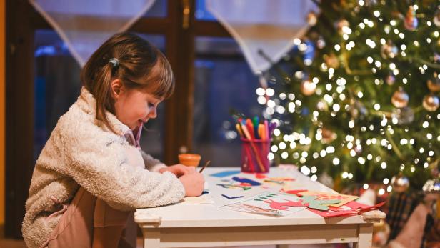Ein Kind bastelt am Tisch mit buntem Papier und Stiften, im Hintergrund steht ein leuchtender Weihnachtsbaum.