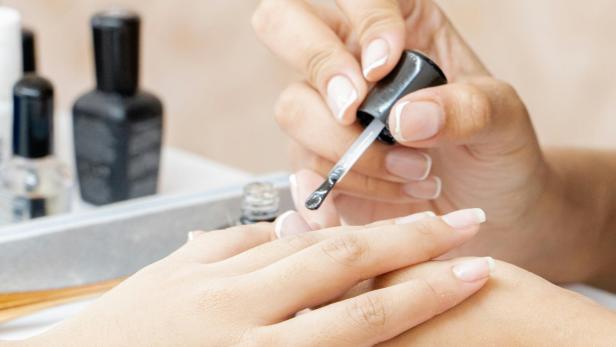 Eine Person trägt in einem Nagelstudio Nagellack auf die Hand einer Kundin auf, im Hintergrund stehen Fläschchen mit Nagellack.