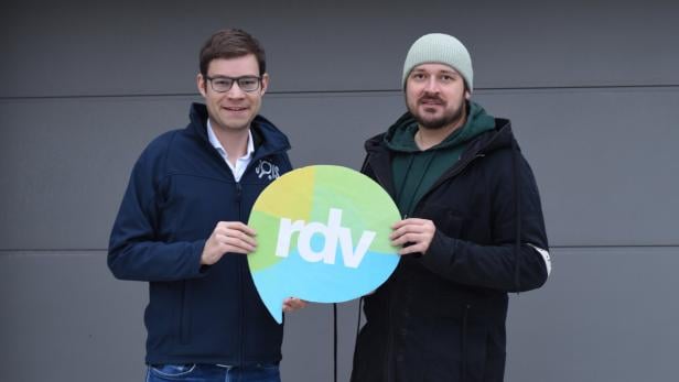 Zwei Männer stehen nebeneinander und halten gemeinsam ein rundes Schild mit der Aufschrift „rdv“.