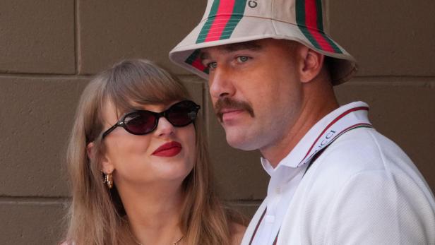 Taylor Swift mit Sonnenbrille und Travis Kelce mit Gucci-Hut schauen einander an.