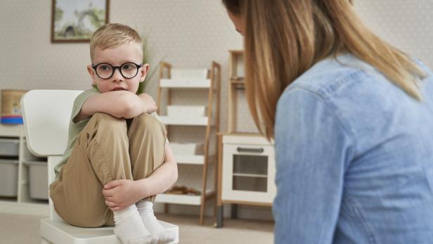 Ein Junge mit Brille sitzt ängstlich auf einem Stuhl, während eine Frau ihm gegenübersteht.