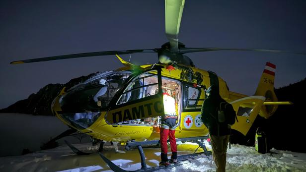 Ein gelber Rettungshubschrauber steht nachts auf verschneitem Boden, zwei Personen stehen beleuchtet daneben.