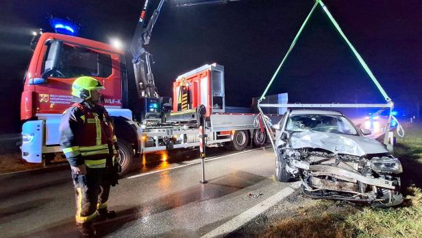 Ein Feuerwehrmann überwacht, wie ein Kran ein stark beschädigtes Auto bei Nacht von der Straße hebt.
