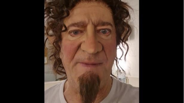 Mann mit Lockenperücke, künstlicher Nase und Bart in einem Raum mit weißer Wand.