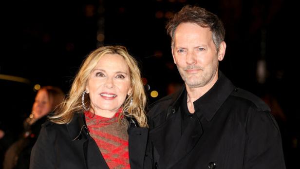 Kim Cattrall und ihr Begleiter tragen schwarze Mäntel vor dunklem Hintergrund.