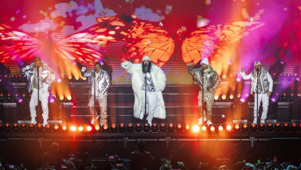 Fünf Sänger in silbernen Outfits stehen auf einer Bühne mit bunten Lichtern und Schmetterlingsprojektion im Hintergrund.